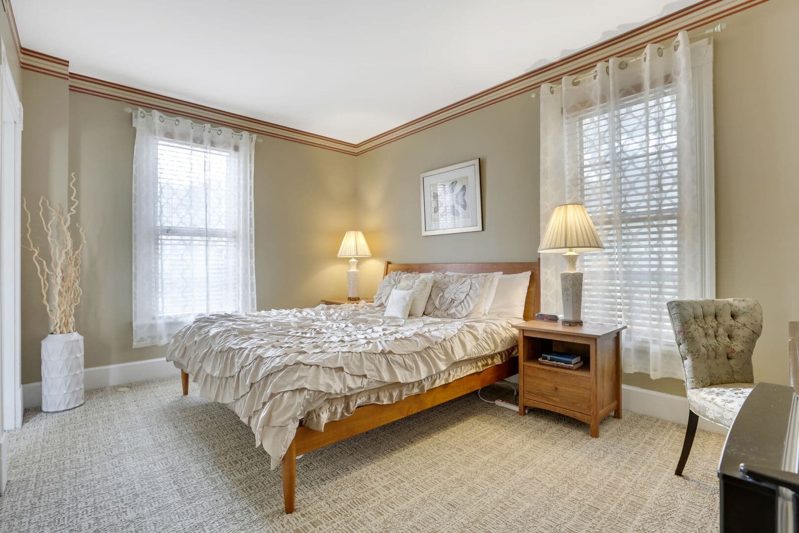 A cozy bedroom with neutral-toned walls and carpet, a wooden bed with ruffled bedding, two windows with sheer curtains, and bedside lamps.