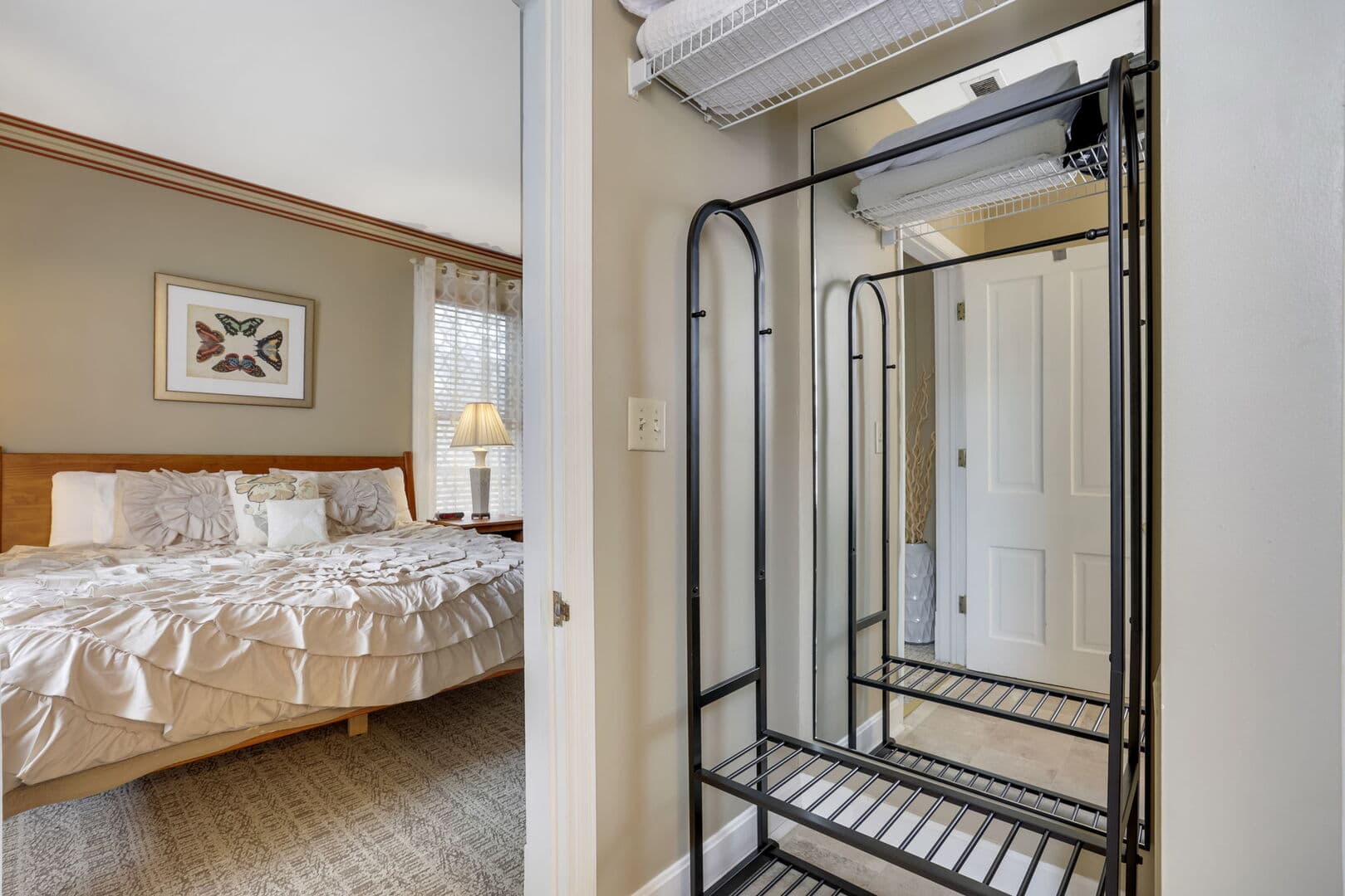 A view into a cozy bedroom with a wooden bed and a mirror, seen from a hallway with a metal rack and a partial reflection.
