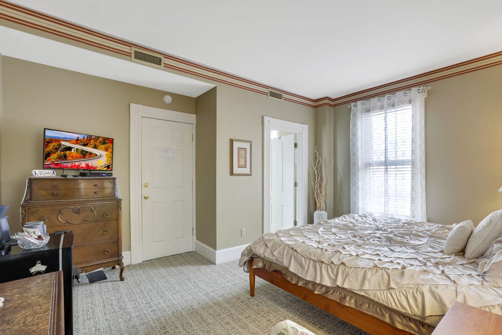 A neutral-toned bedroom with a wooden bed, a large window, and a TV on a dresser.