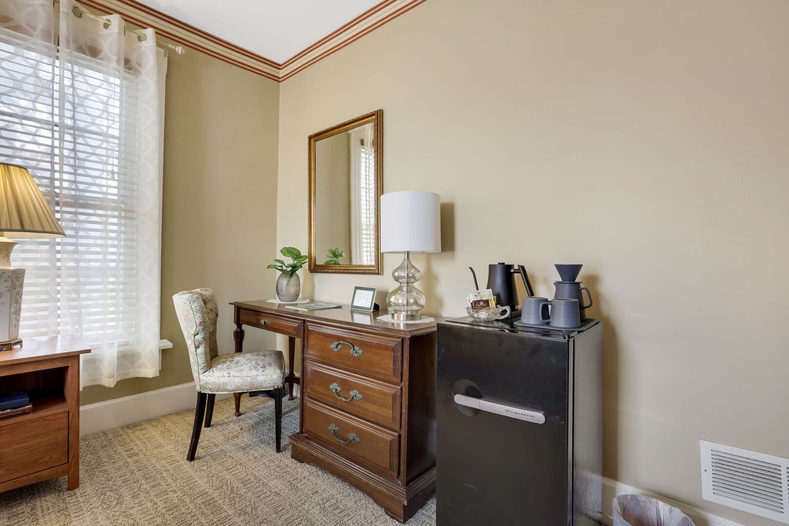 A warm-toned room features a wooden desk with a mirror, a chair, a mini-fridge, a coffee station, and a window with blinds.