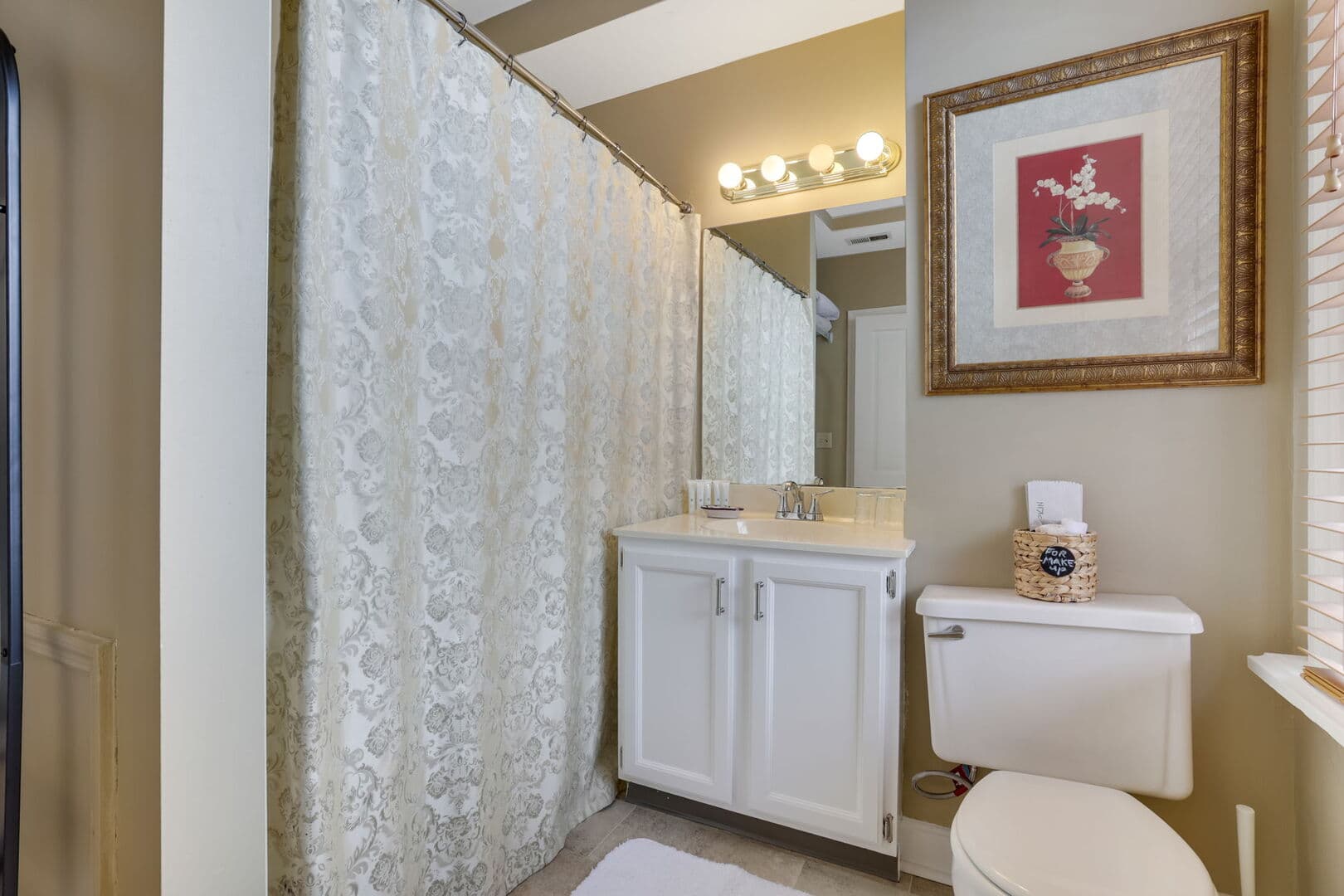 A well-lit bathroom features a white vanity, a mirror, a toilet, a patterned shower curtain, and a framed floral print on the wall.