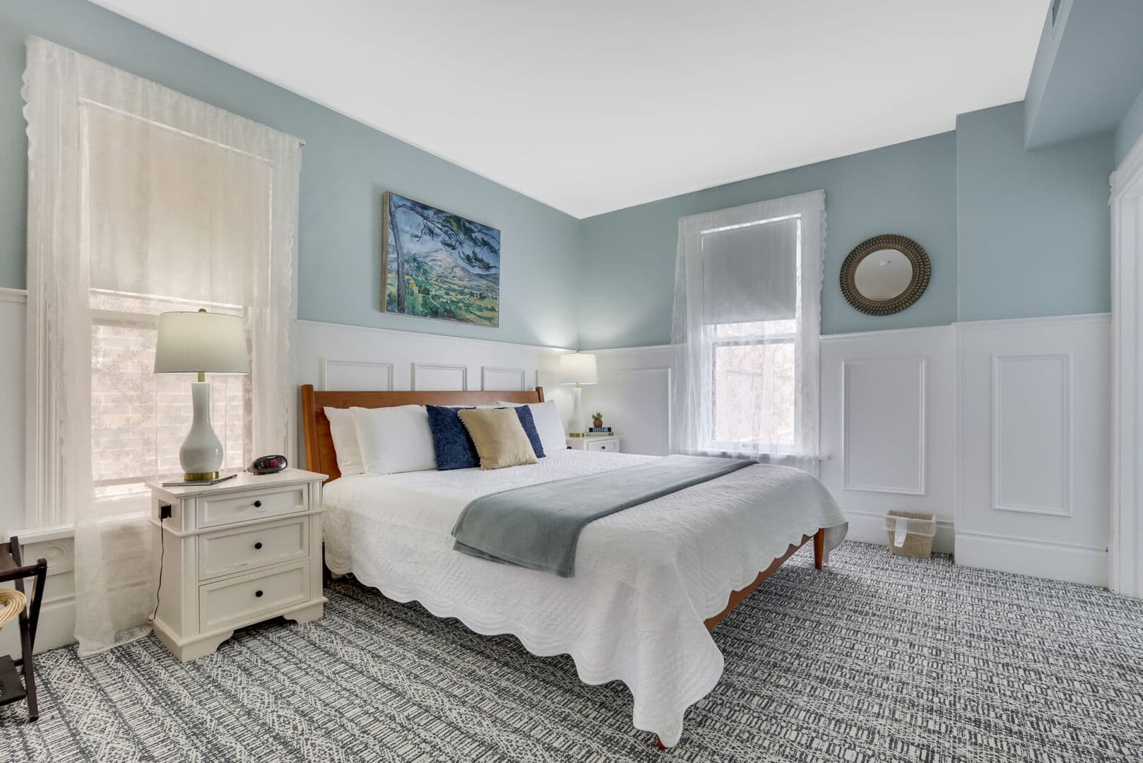 A spacious bedroom with light blue walls and white wainscoting, featuring a queen bed, two windows, and bedside tables with lamps.