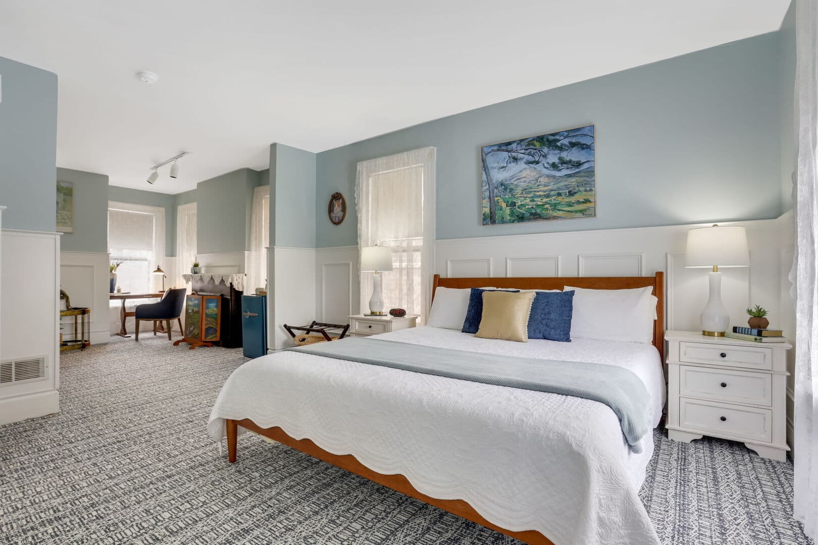 A serene bedroom with a queen bed, light blue walls, white wainscoting, and a distinct seating area with a table and chairs in the background.