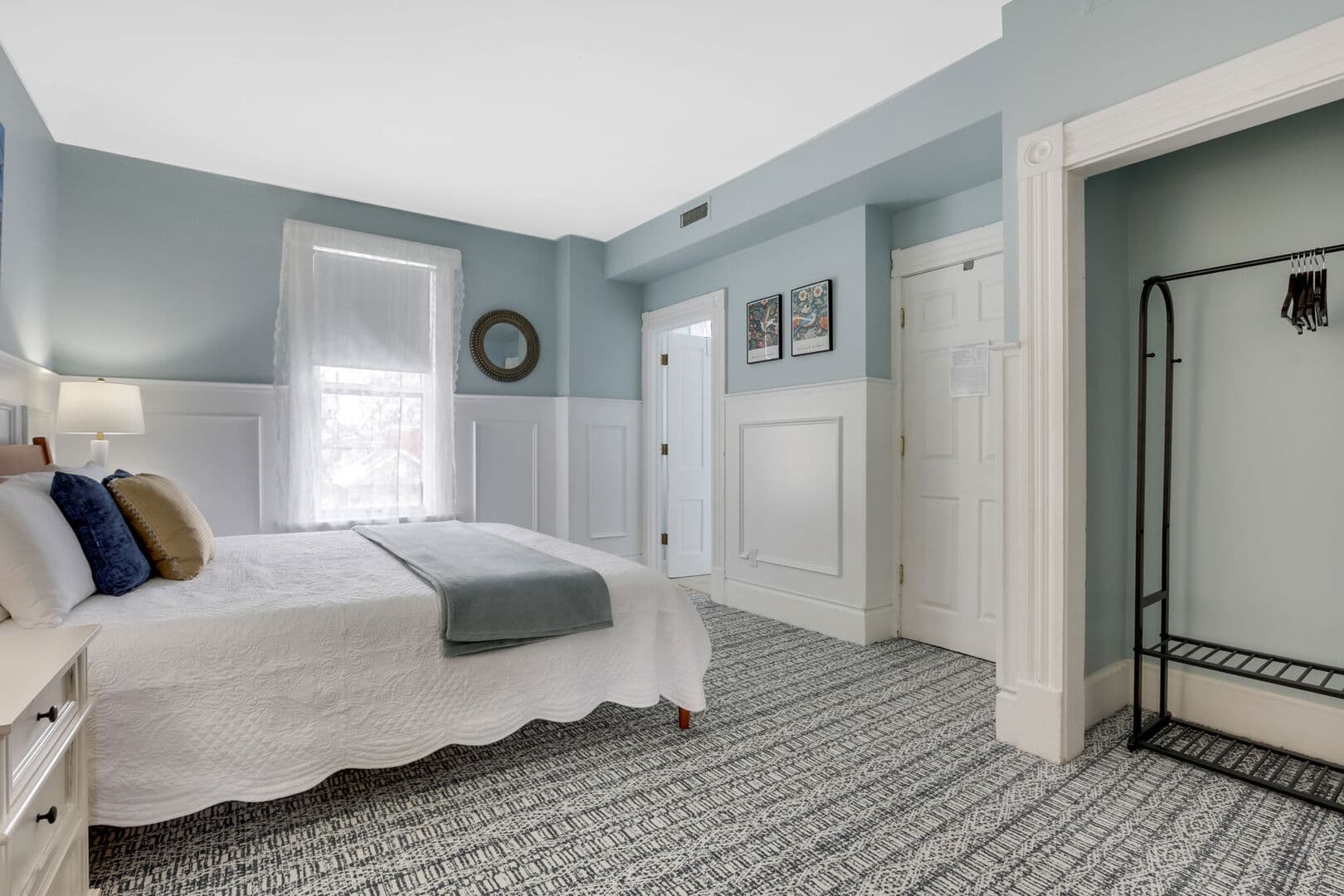 A bright bedroom with light blue walls and white wainscoting features a queen bed, two windows, and an open closet rack.
