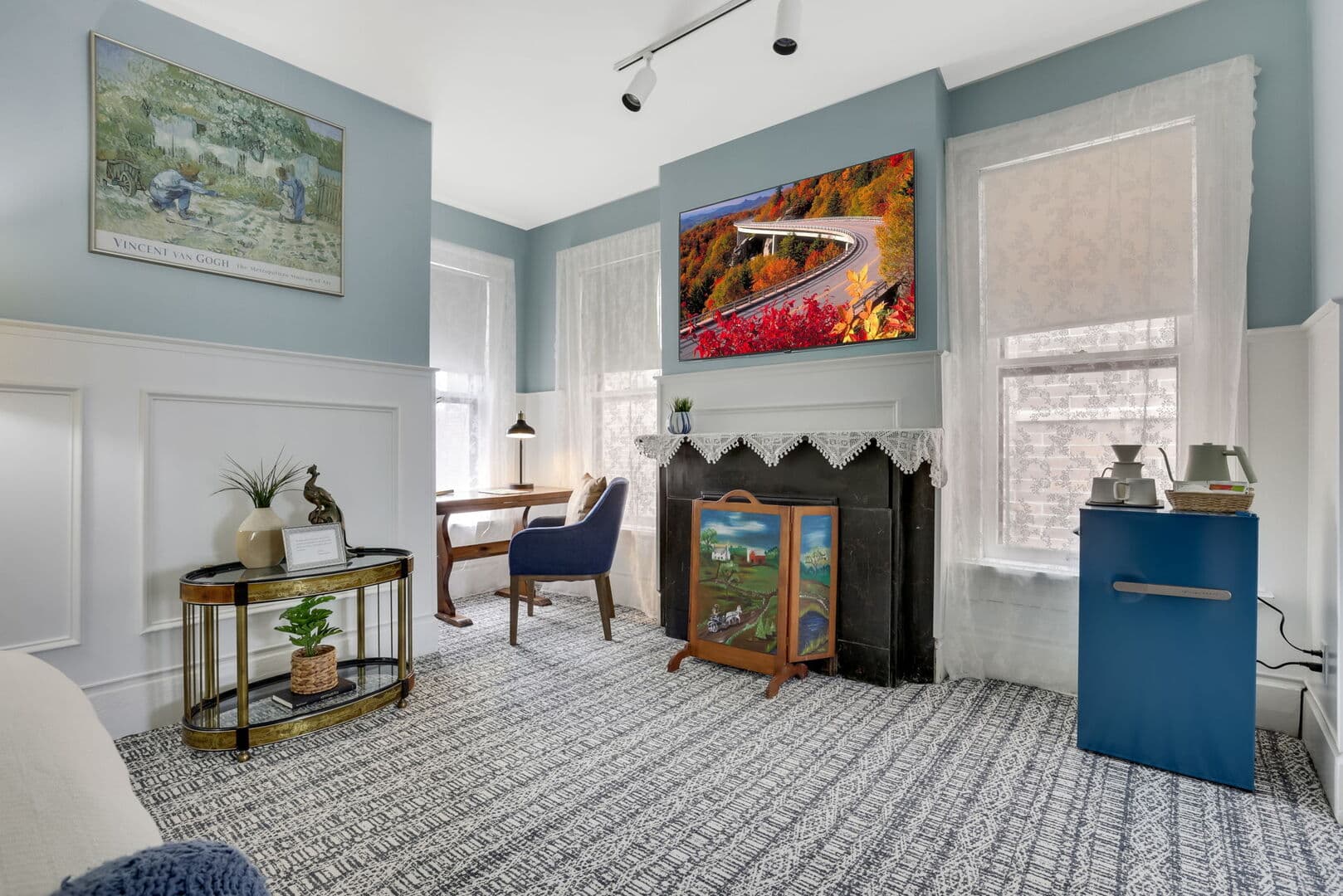 A vibrant living space with light blue walls, white wainscoting, and a patterned rug. Features include a TV, a desk, and decorative fire screen..