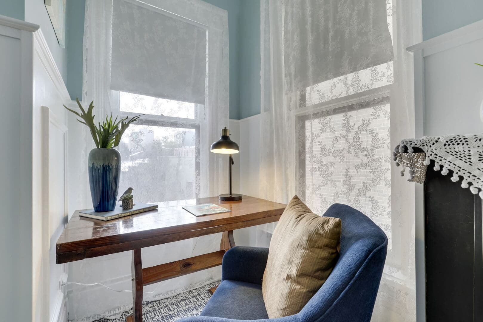 A cozy nook with light blue walls and white wainscoting features a wooden desk, a blue armchair, a lamp, and two windows with patterned blinds.