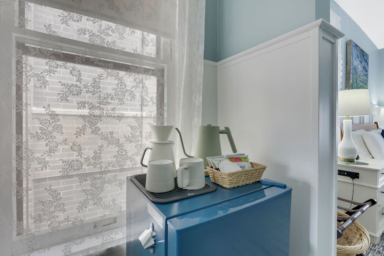 A bright corner with light blue walls and white wainscoting features a blue mini-fridge with a coffee setup, next to a window with blinds.
