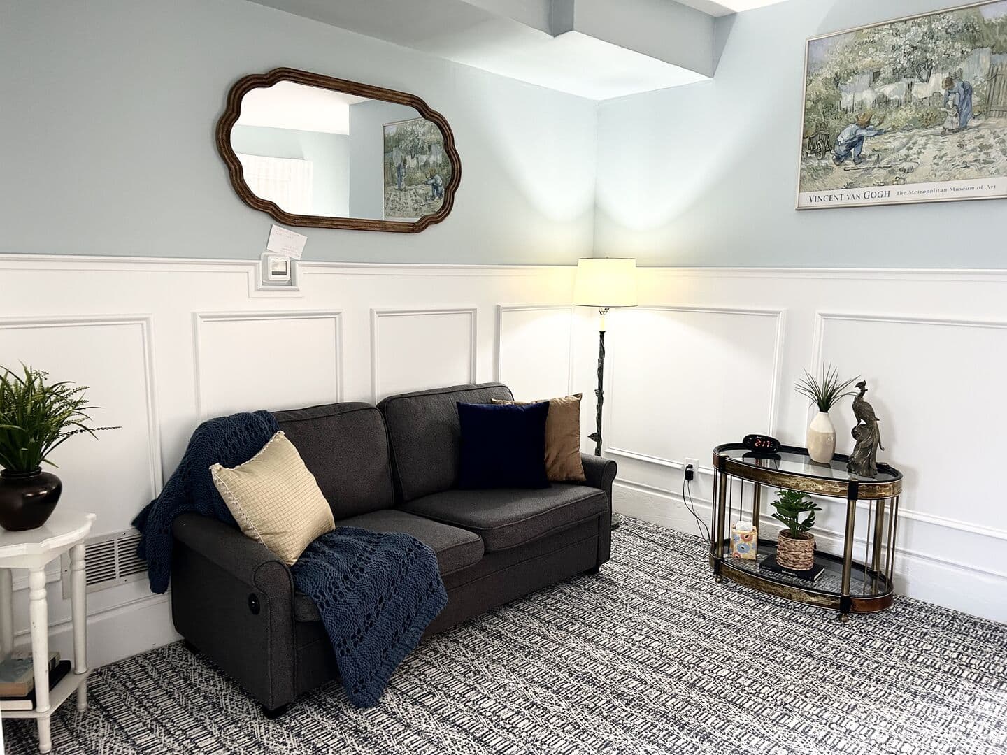 A cozy living area with light blue walls and white wainscoting features a dark sofa with throws, a decorative mirror, and a side table.