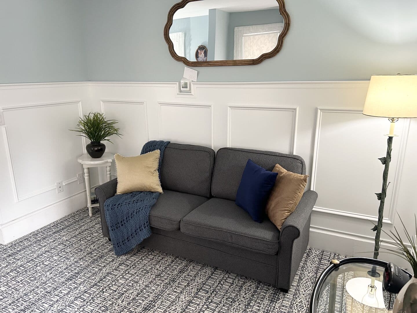 A welcoming sitting area with light blue walls and white wainscoting, featuring a gray sofa with blue and tan pillows, and a unique mirror.