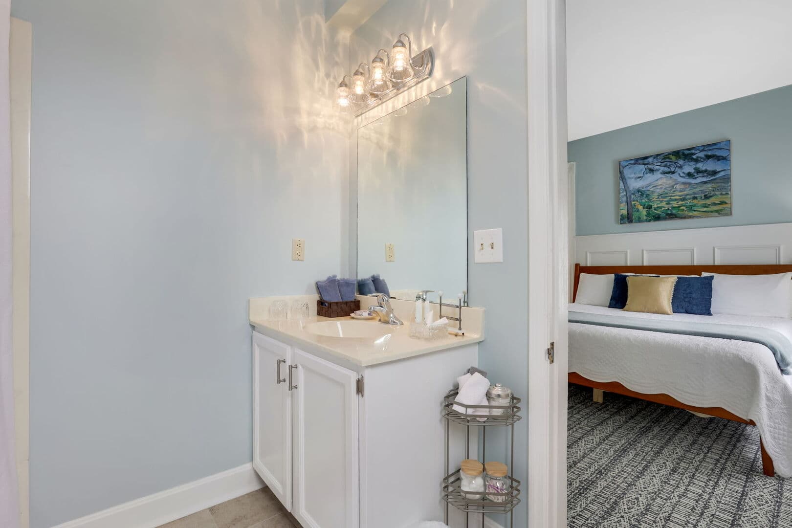 A well-lit vanity area with a white cabinet, mirror, and light blue walls, adjacent to a bedroom with a visible bed and artwork.