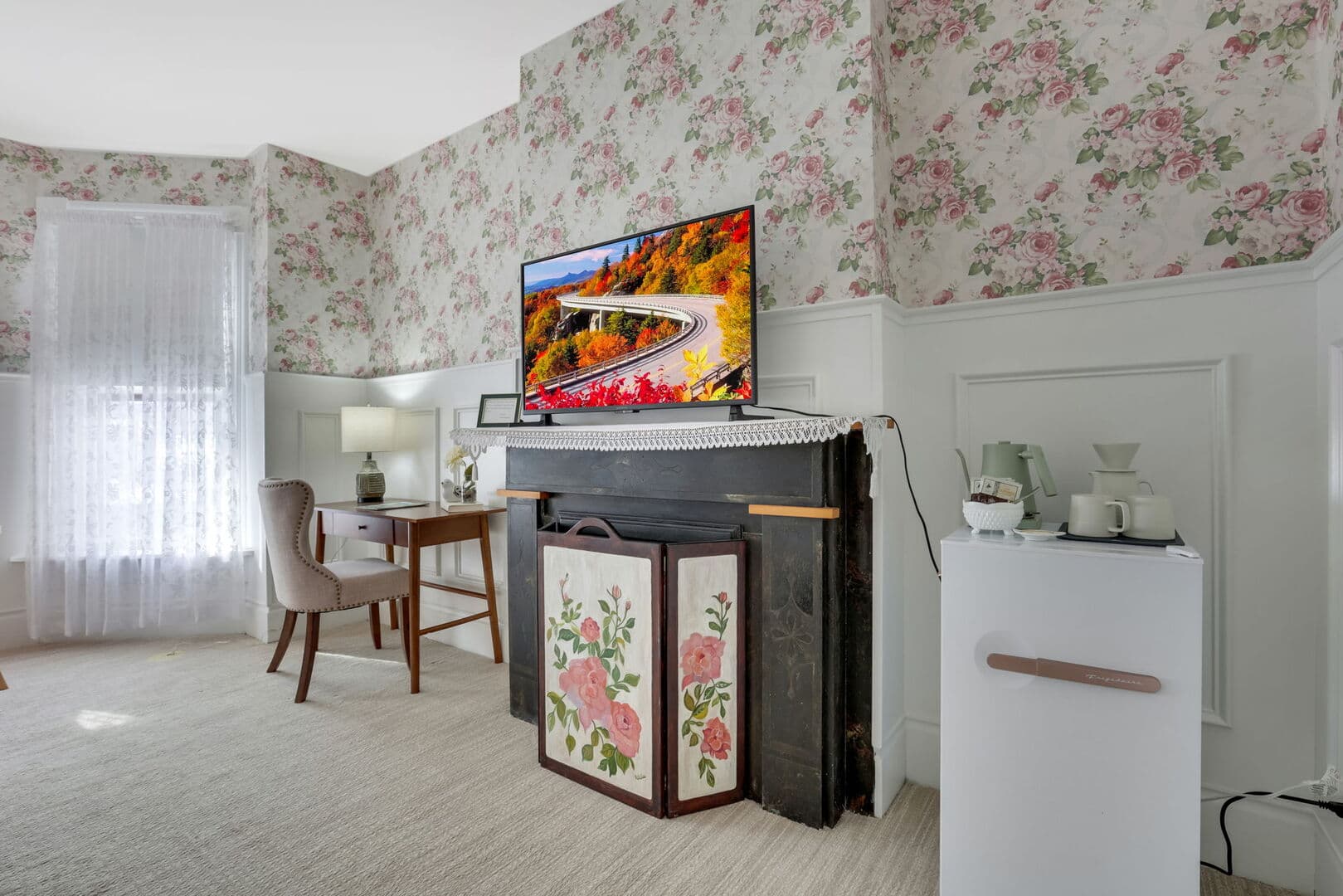 A floral-wallpapered room with white wainscoting, featuring a TV above a decorative fireplace, a desk with a chair, and a mini-fridge.