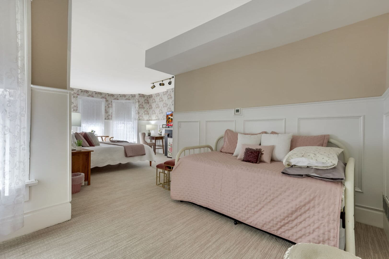 A spacious, light-filled room with a daybed, soft pink bedding, neutral walls, white wainscoting, and a curved window area.