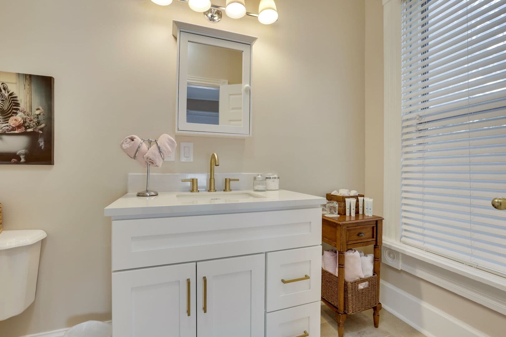 A brightly lit bathroom features a white vanity with gold accents, a medicine cabinet, a toilet, and a window with blinds.