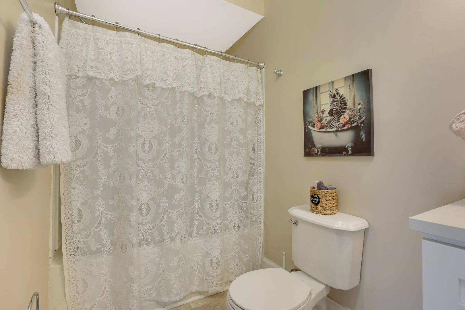 A pristine bathroom features a white toilet, a patterned shower curtain, a towel hanging on a rod, and a framed picture above the toilet.