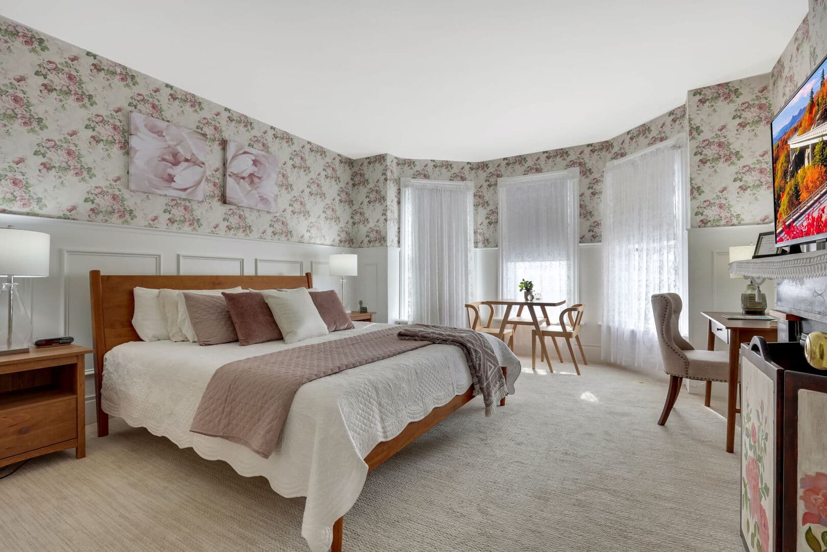 A charming bedroom with floral wallpaper and white wainscoting, featuring a wooden bed, a small dining area, and a desk.