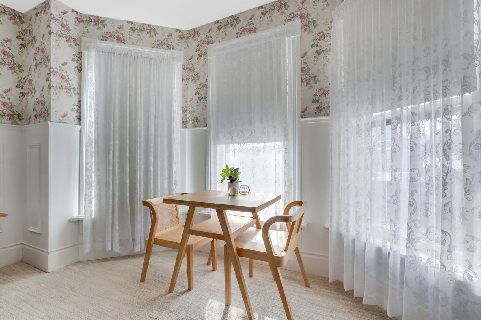 A bright dining nook with floral wallpaper and white wainscoting, featuring a light wood table, two chairs, and sheer window coverings.