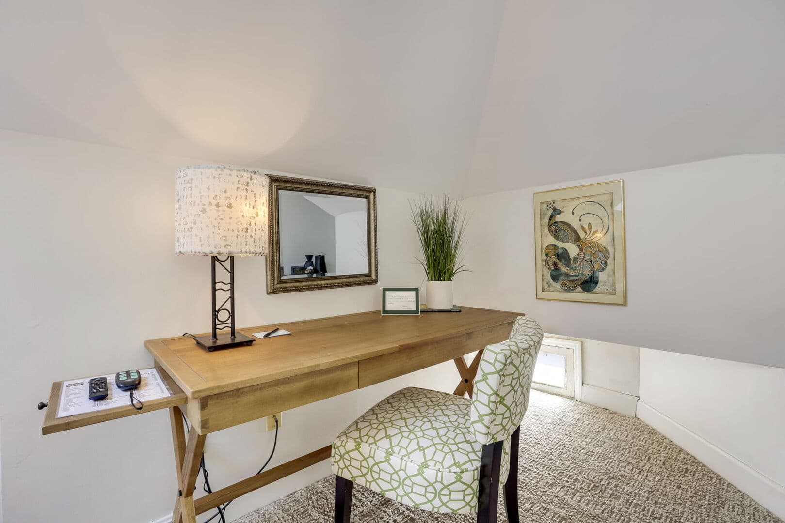 A bright workspace with a sloped ceiling, featuring a wooden desk, a patterned chair, a mirror, and a lamp.