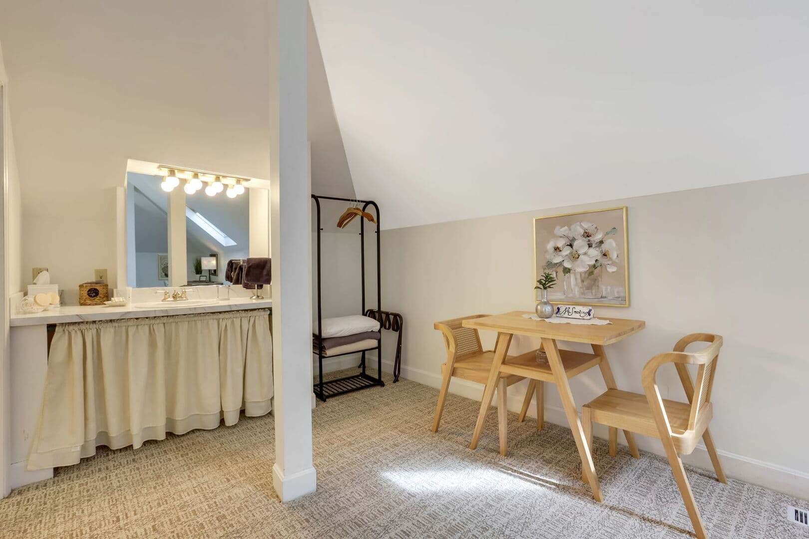 A spacious third-floor room with sloped ceilings, featuring a dining area with a round table and chairs, and a vanity area with a mirror.