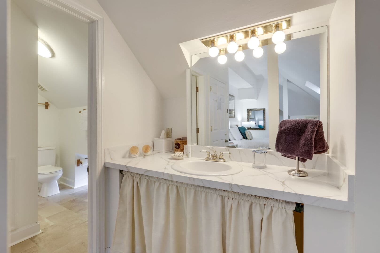 A brightly lit bathroom vanity with a white counter and skirted cabinet, featuring a mirror, sink, and a view into another bathroom area.