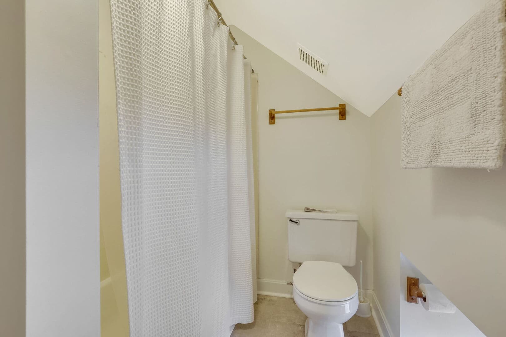 A small, bright bathroom with a sloped ceiling, featuring a white toilet, a shower with a white curtain, and two towel racks.
