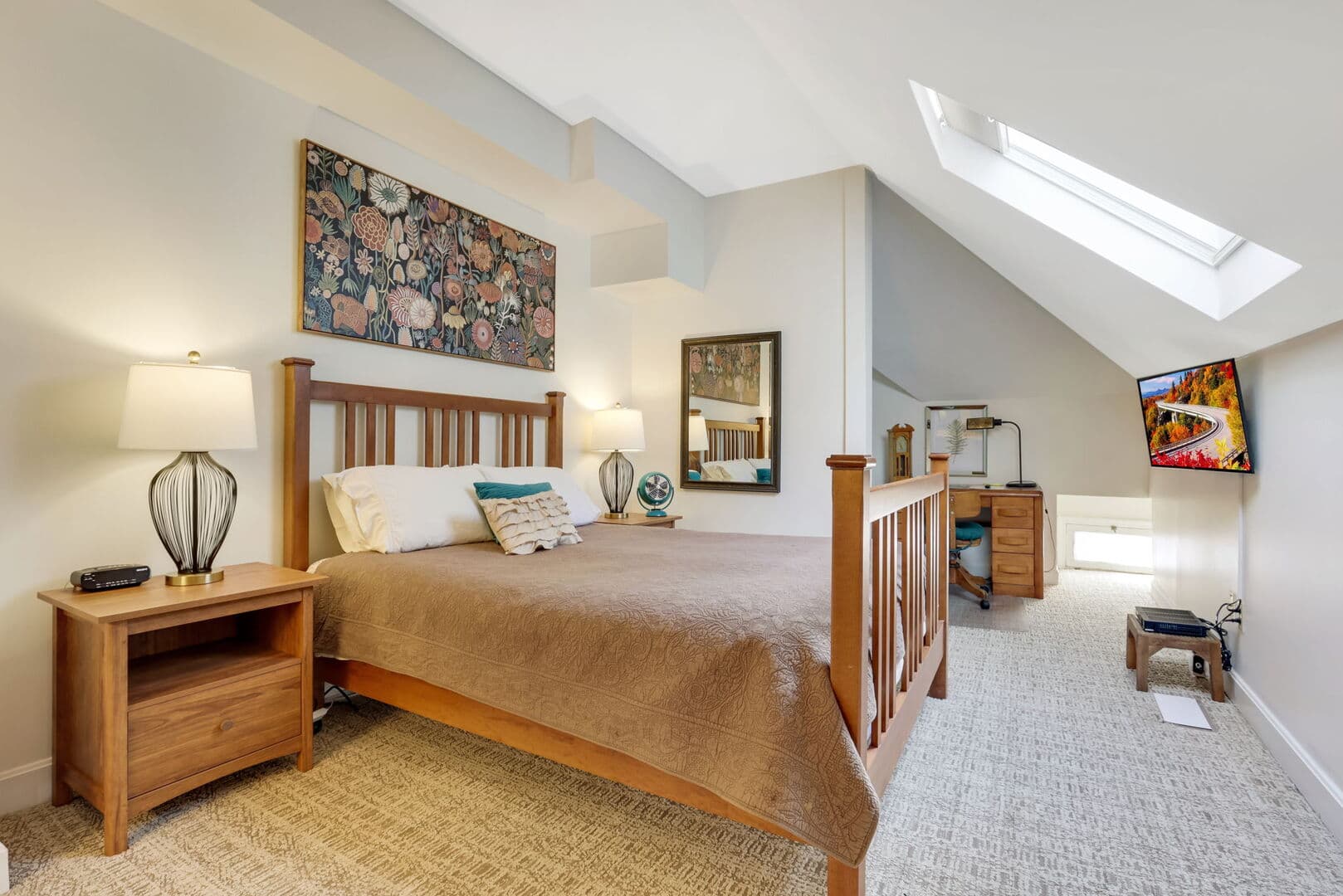 A spacious bedroom with a wooden queen bed, patterned art, a bedside table, a skylight, and a TV on a sloped wall.