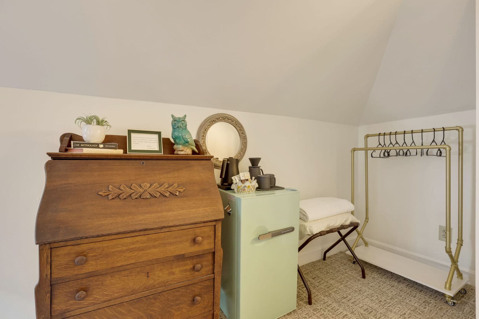 A cozy space features a vintage wooden desk, a pastel green mini-fridge, and a gold clothing rack, all under a sloped ceiling.