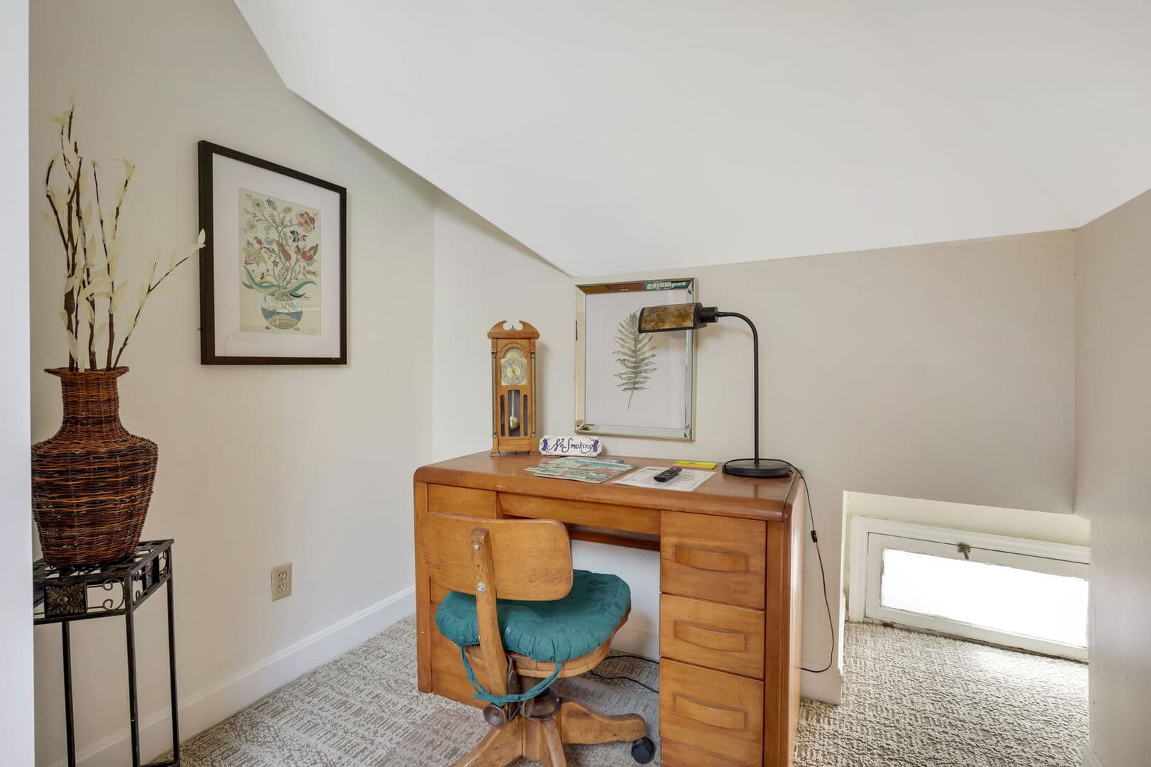 A functional and inviting workspace with a wooden desk, a teal chair, a lamp, framed art, and a decorative vase, under a sloped ceiling.