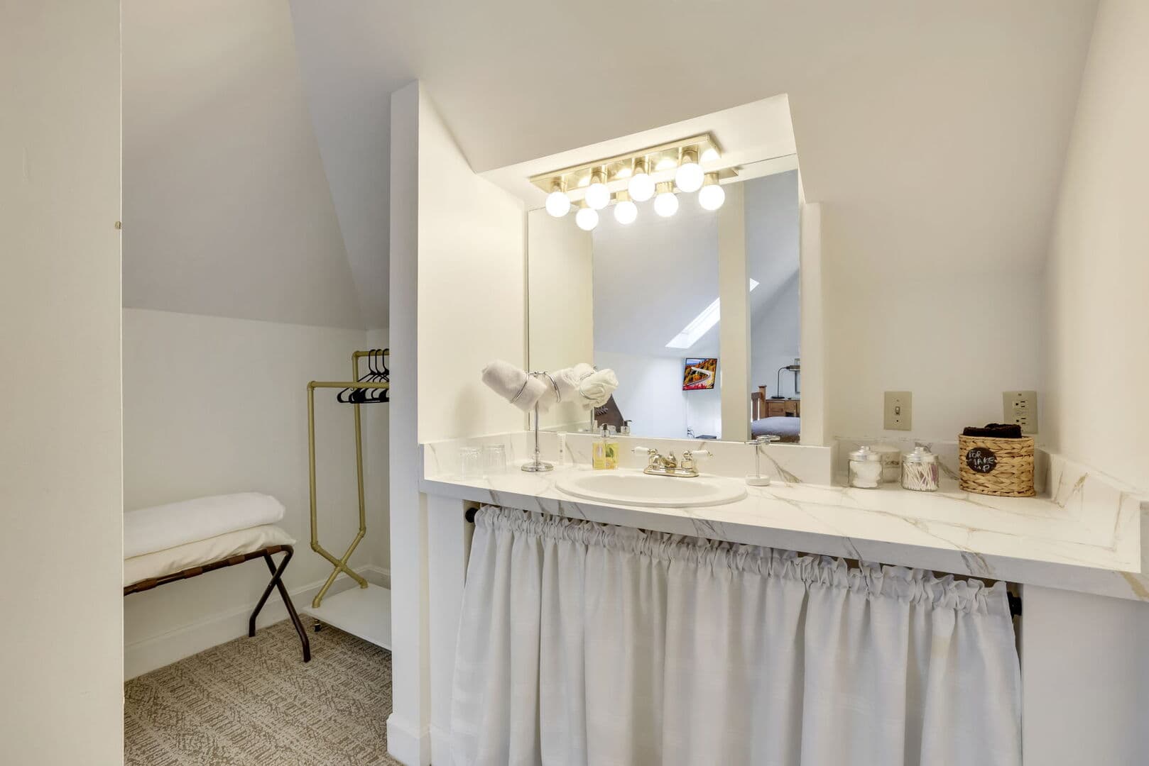 A well-lit bathroom vanity with a white counter and skirted cabinet, a large mirror, and a small seating area.