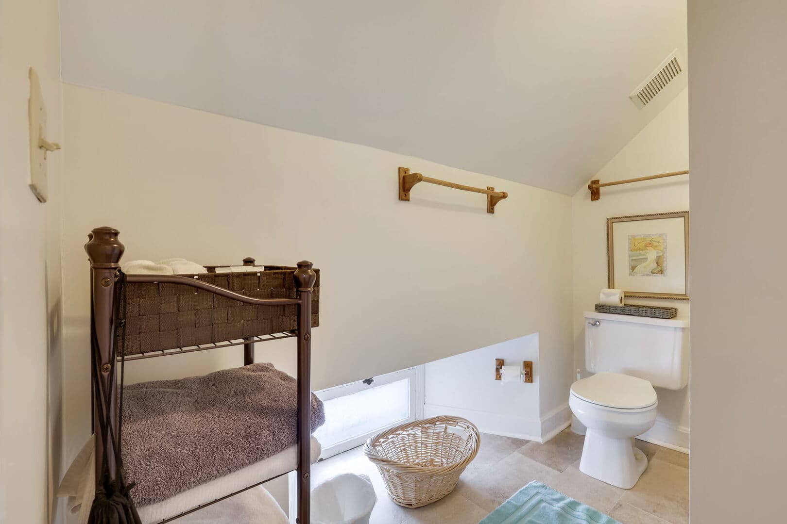 A quaint bathroom/dressing area with a sloped ceiling, featuring a toilet, a small changing station with a basket, and towel racks.