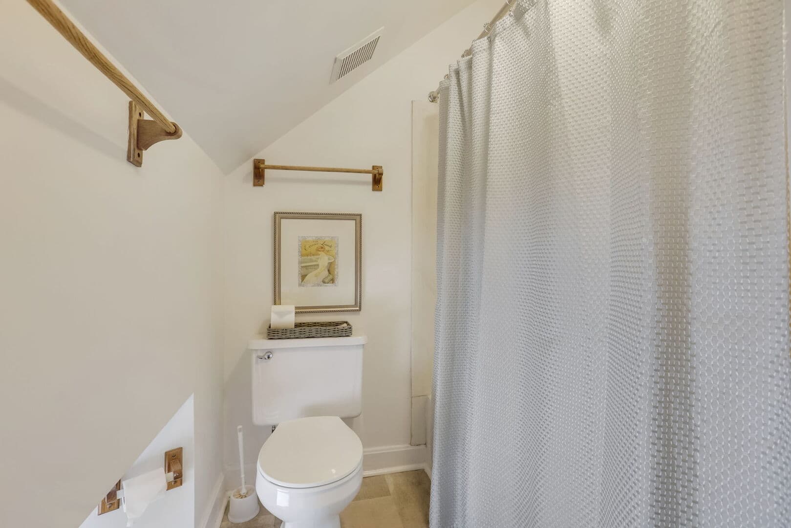 A cozy third-floor bathroom with a sloped ceiling, featuring a toilet, a shower with a light-colored curtain, and framed artwork.