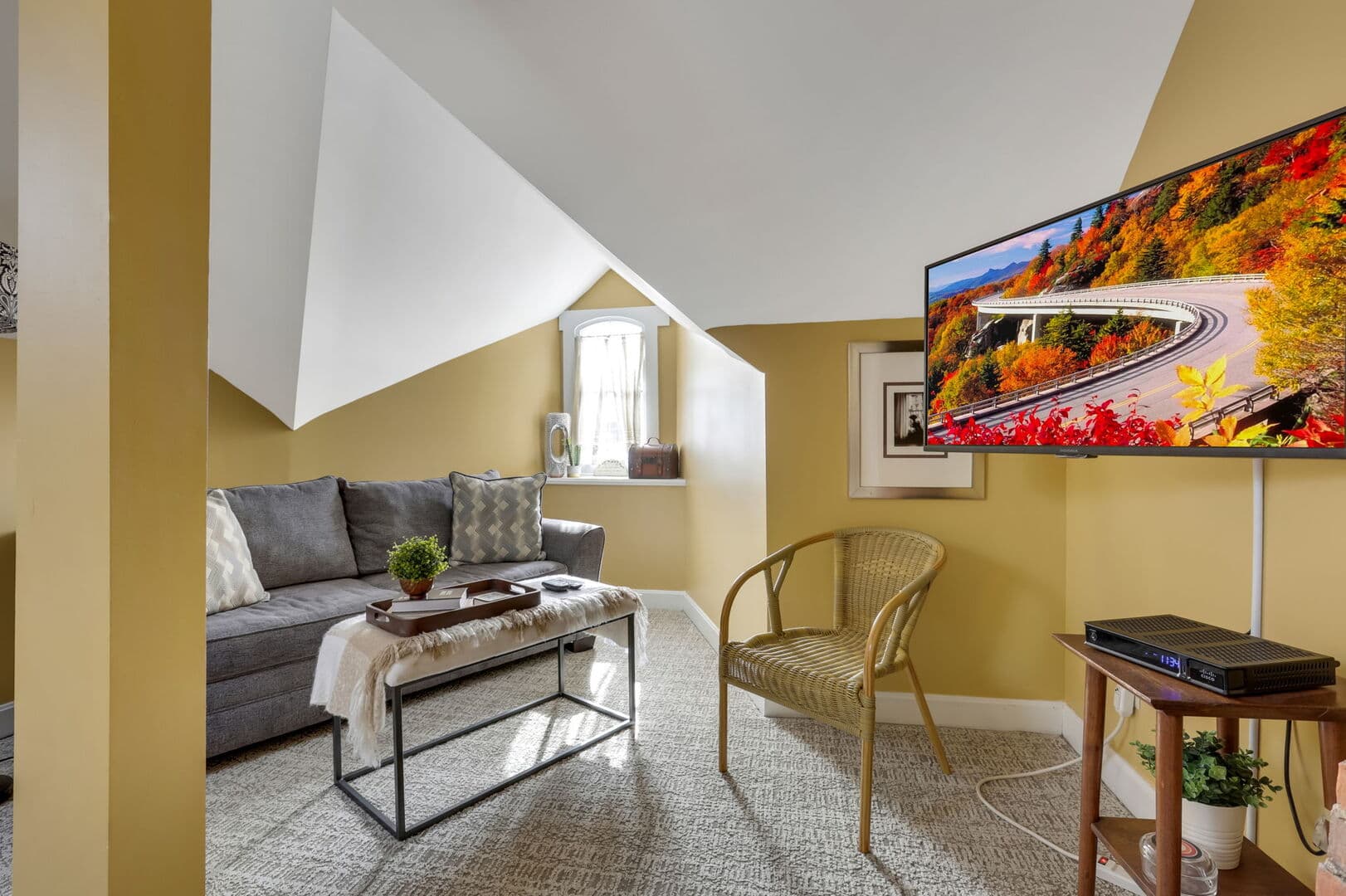 A cozy living area with golden walls, a sloped ceiling, a gray sofa, a coffee table, a TV, and a charming window nook.