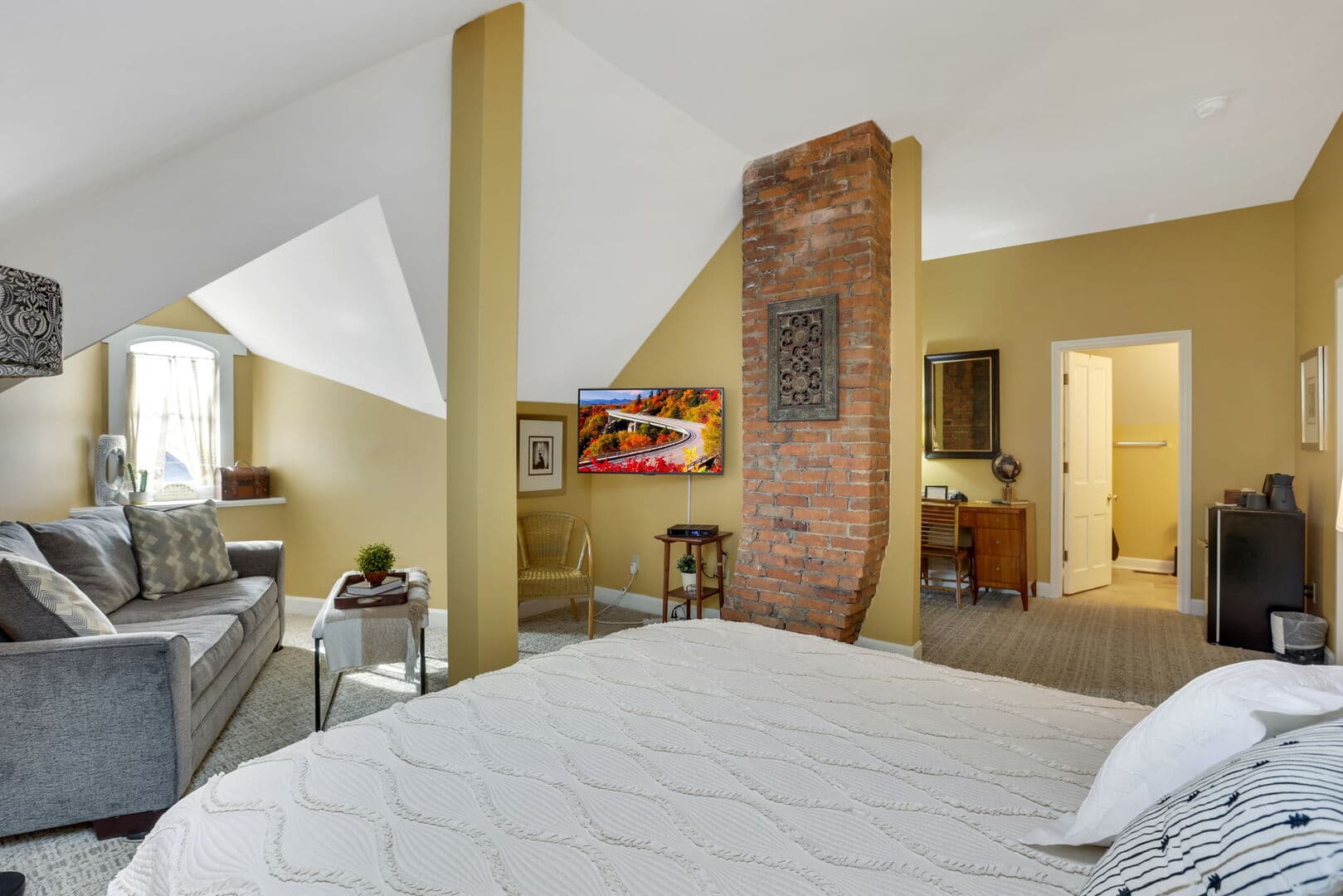 A spacious third-floor studio with golden walls, a queen bed, a living area with a sofa and TV,  a brick accent wall and doorway leading to a bathroom.