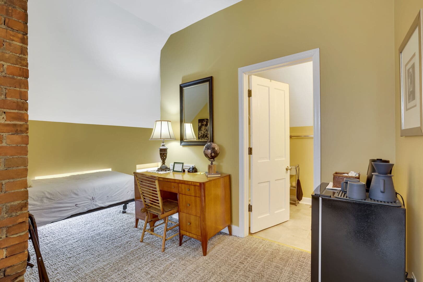 A golden-walled third-floor room with a twin bed, a wooden desk with a mirror, a lamp, and a mini-fridge near an open doorway.