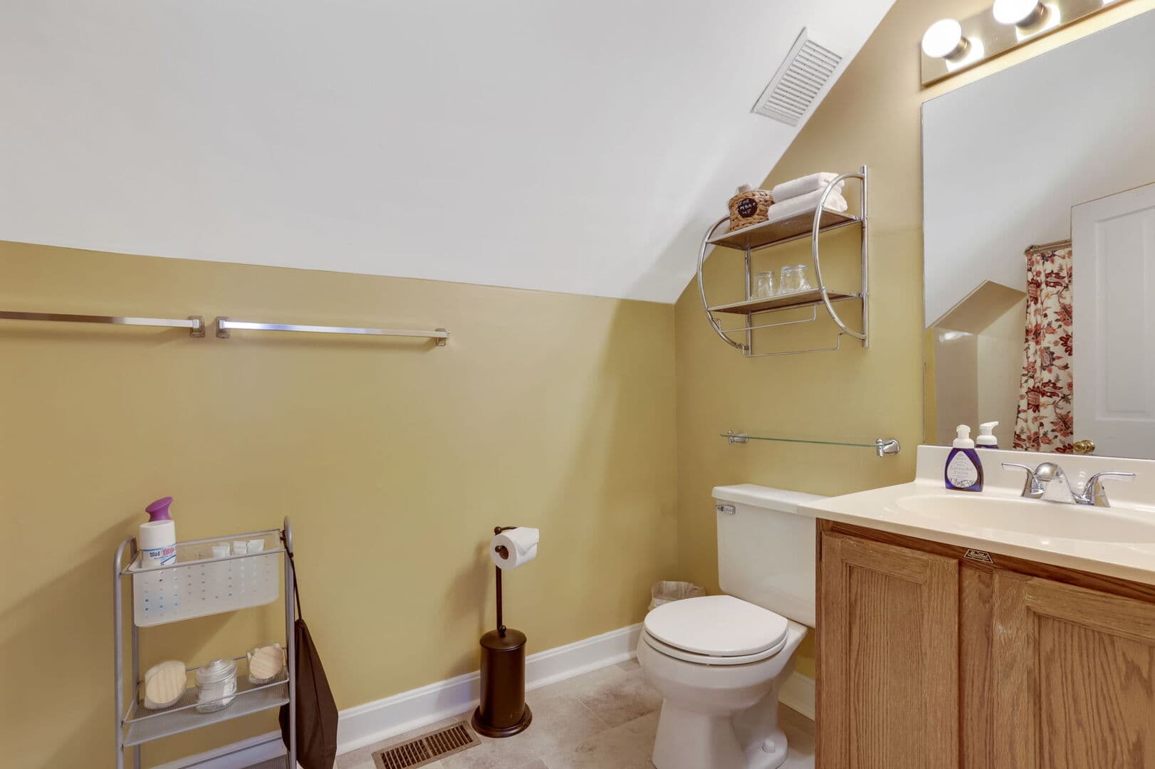 A warm-toned bathroom with a sloped ceiling, featuring a toilet, a wooden vanity with a mirror, and metal shelving units.