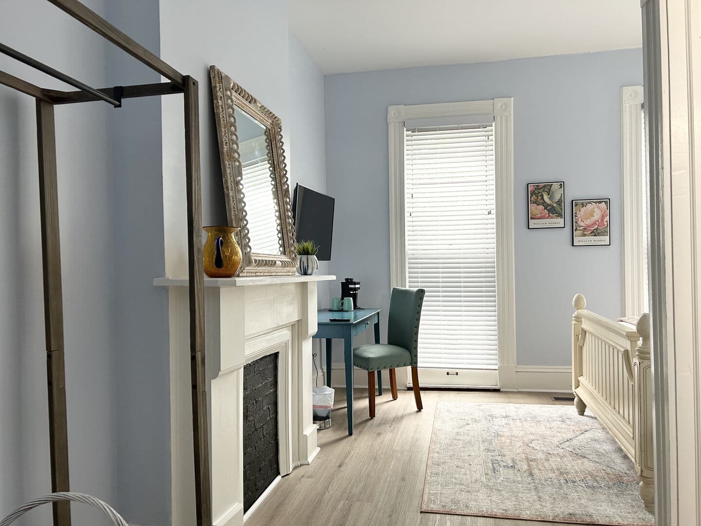 A bedroom with light blue walls features a decorative fireplace, a large mirror, a TV, a desk with a teal chair, and a glimpse of a bed.