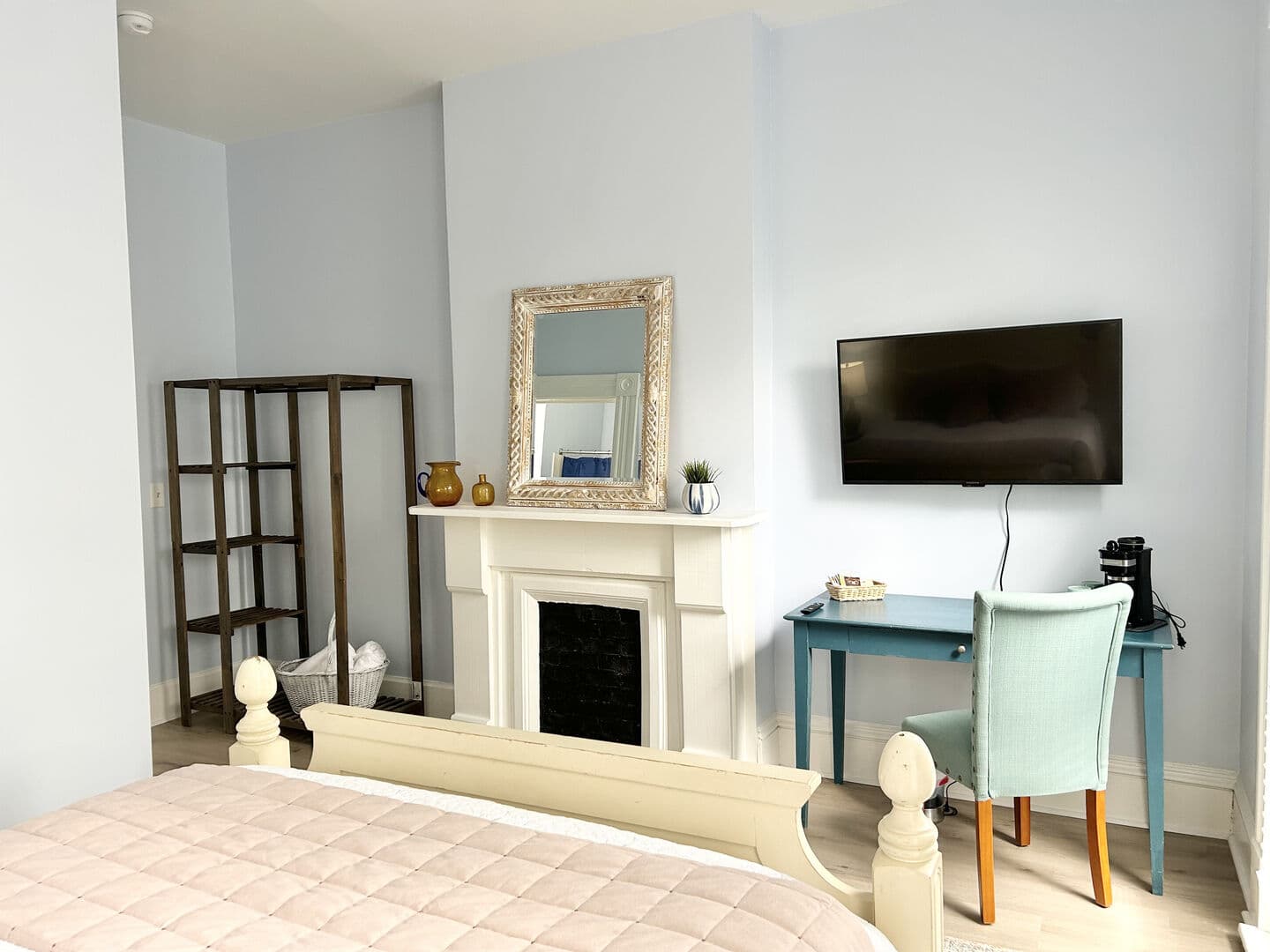 A serene bedroom with light blue walls features a cream bed, a decorative fireplace with a mirror, a TV, a blue desk, and a teal chair.