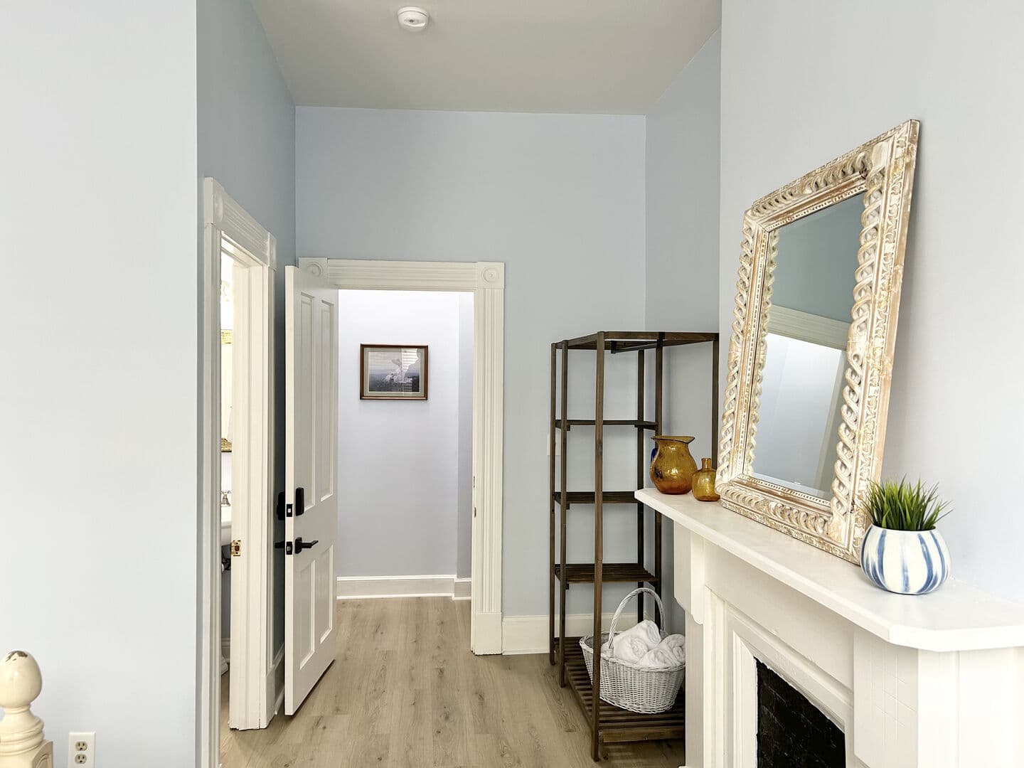 A bright hallway with light blue walls, light wood flooring, a decorative fireplace with a mirror, and an open doorway leading to another room.