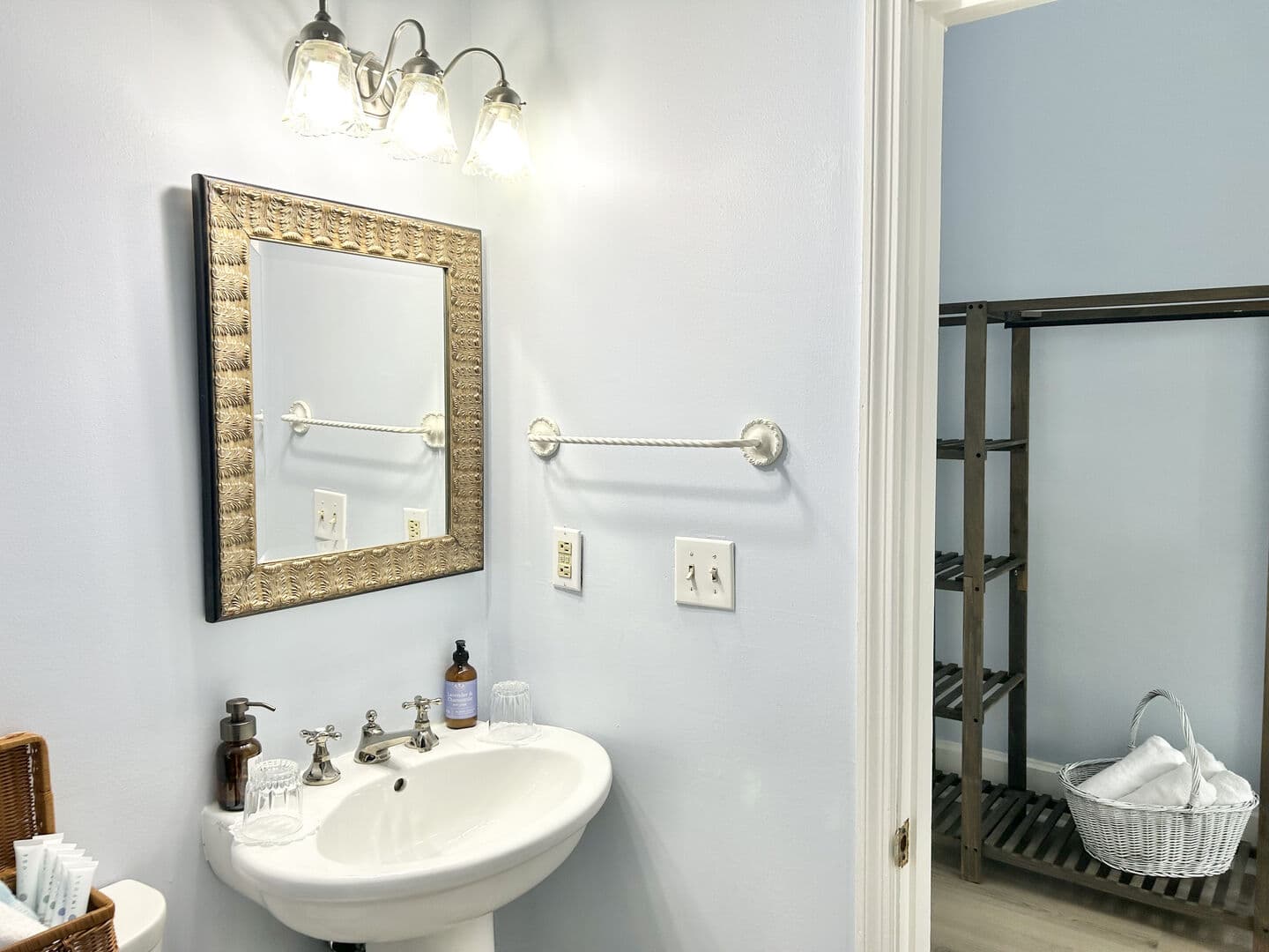 A clean bathroom with light blue walls features a pedestal sink, a framed mirror, a towel bar, and a glimpse of a nearby shelving unit.