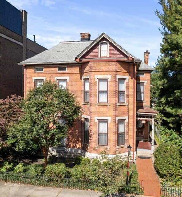 A large, historic brick house with multiple windows, a grey roof, and lush green landscaping, viewed from the front.