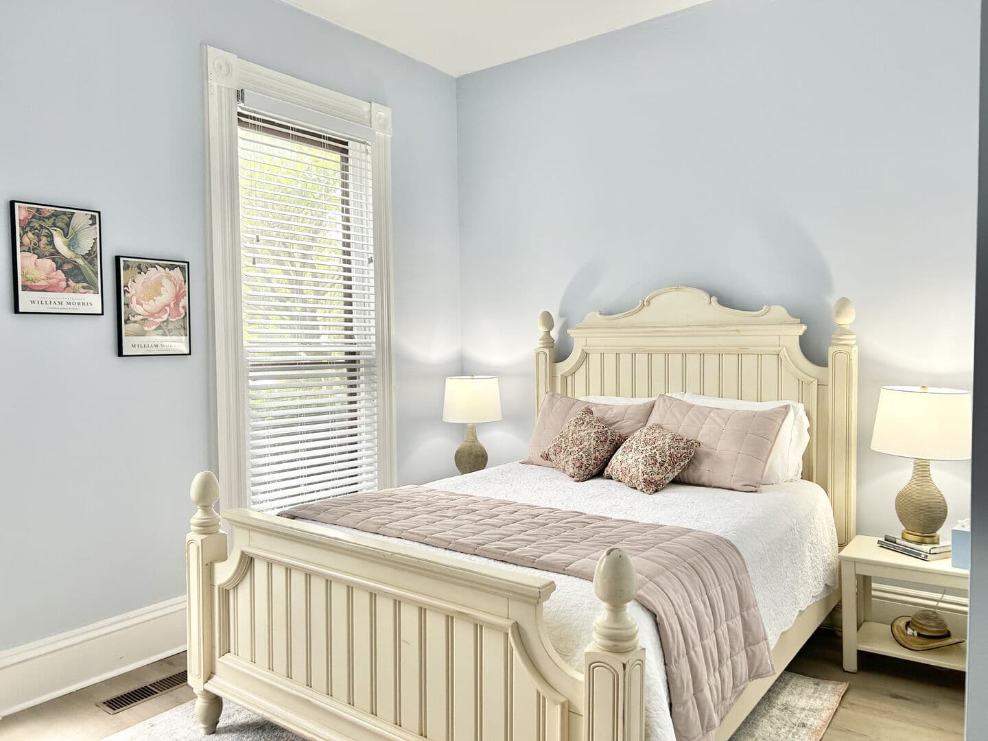 A cozy bedroom with light blue walls, a white wooden bed with a pink throw, two lamps on bedside tables, and framed artwork beside a window.
