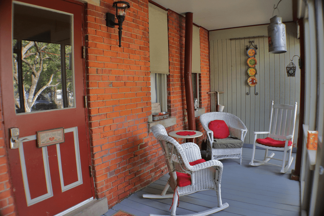 Front porch of a brick house featuring white wicker patio furniture with red seat cushions