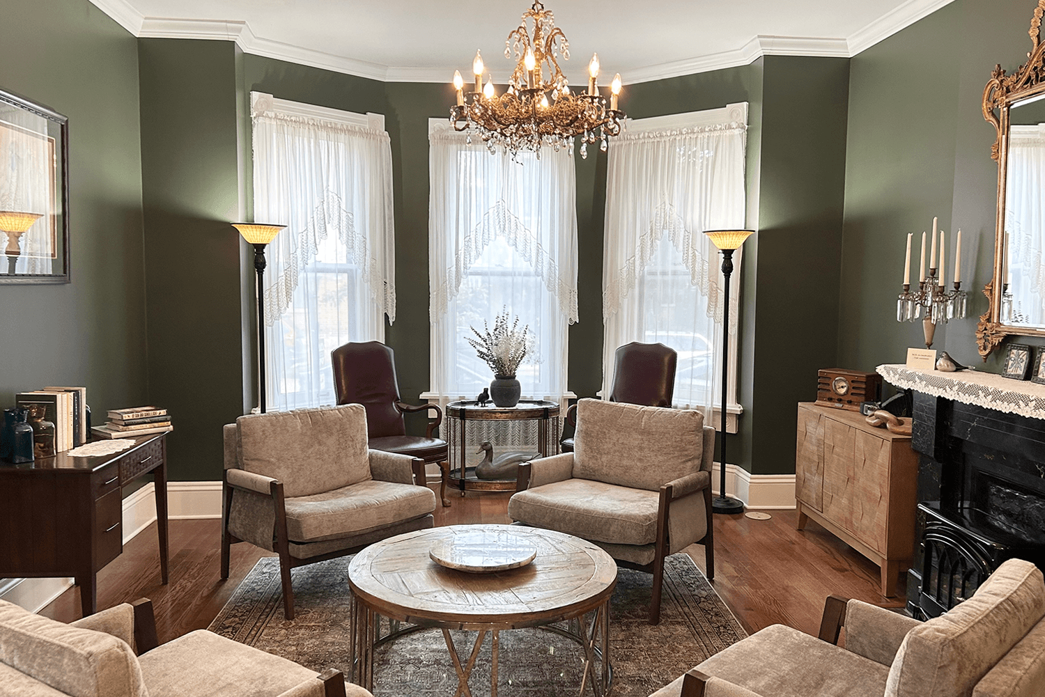 A cozy living room with plush seating, a round coffee table, large windows with lace curtains, and decorative lighting.