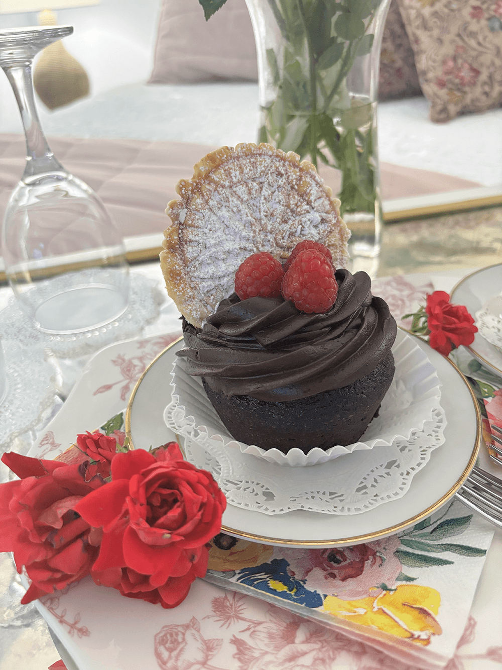 A decadent chocolate cupcake topped with raspberries and a cookie, surrounded by red roses on a floral plate.