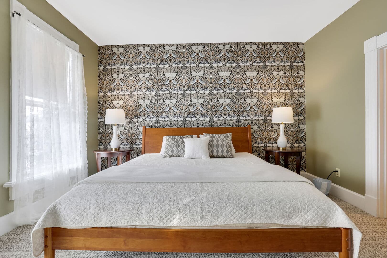 A stylish bedroom with an olive green accent wall featuring patterned wallpaper, a wooden queen bed, white bedding, and two bedside lamps.