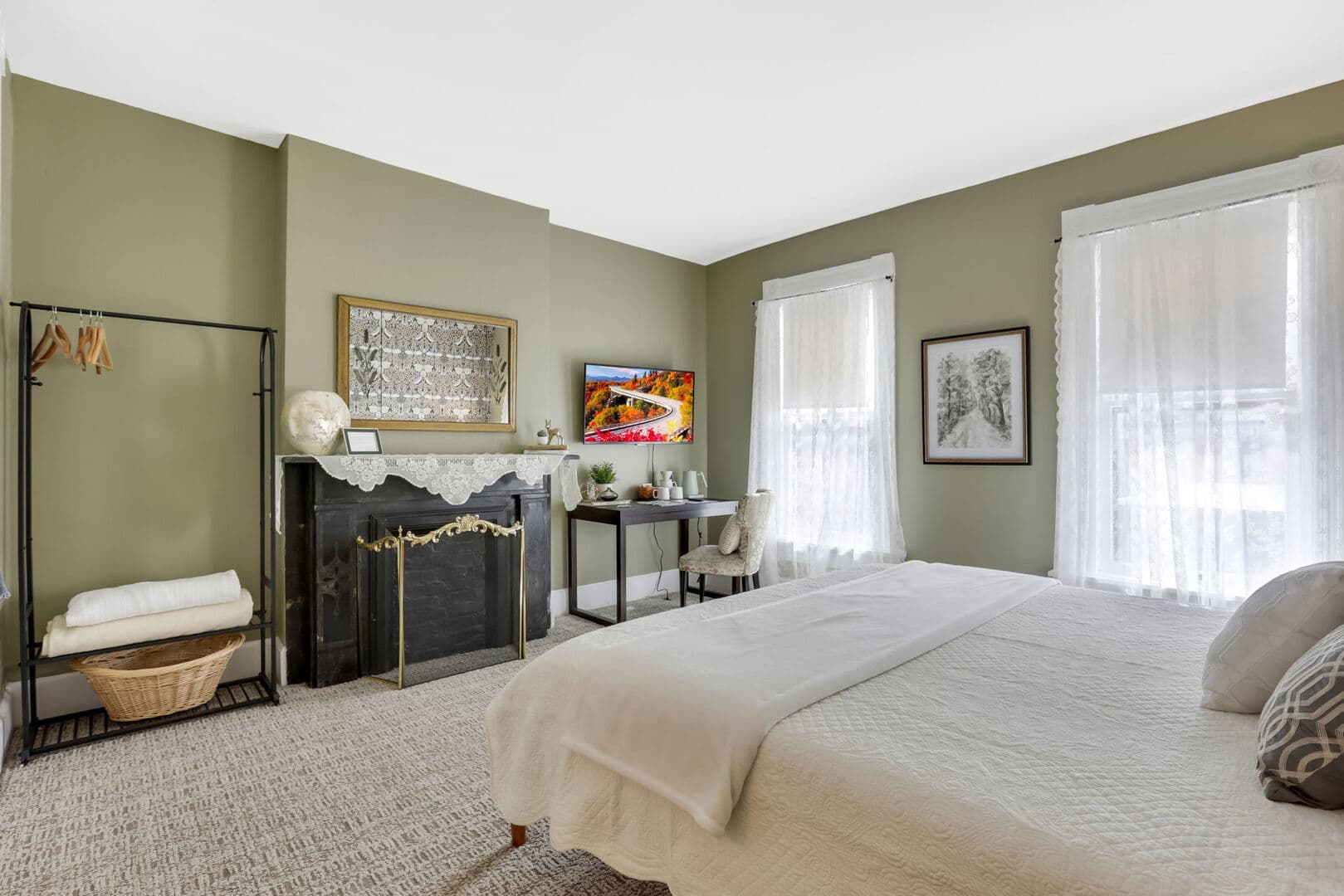 A spacious bedroom with olive green walls, a queen bed, a fireplace with a TV above it, a desk, and a clothing rack.