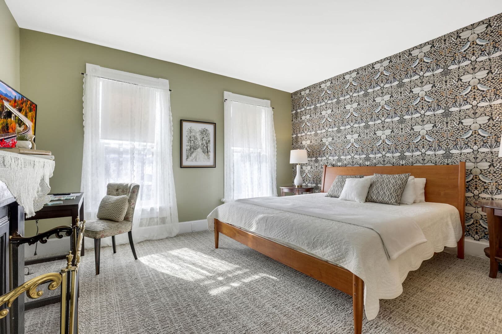A bedroom with an accent wall of patterned wallpaper and olive green walls, featuring a wooden queen bed, two windows, and a desk area.