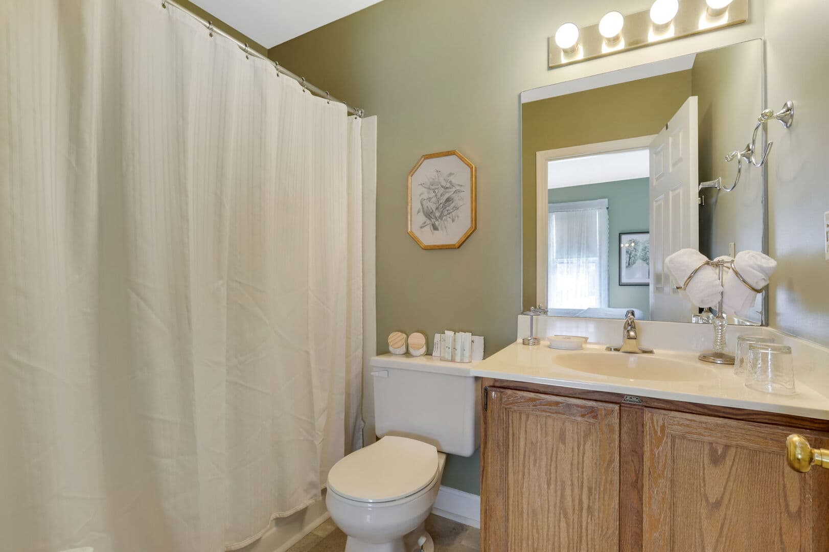 A charming bathroom with olive green walls, a wooden vanity, a toilet, a white shower curtain, and a framed botanical print.