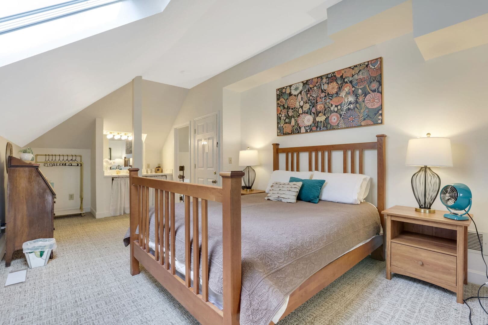 A bright bedroom with a wooden bed, patterned artwork, a bedside table with a fan, and a glimpse of an en-suite bathroom.