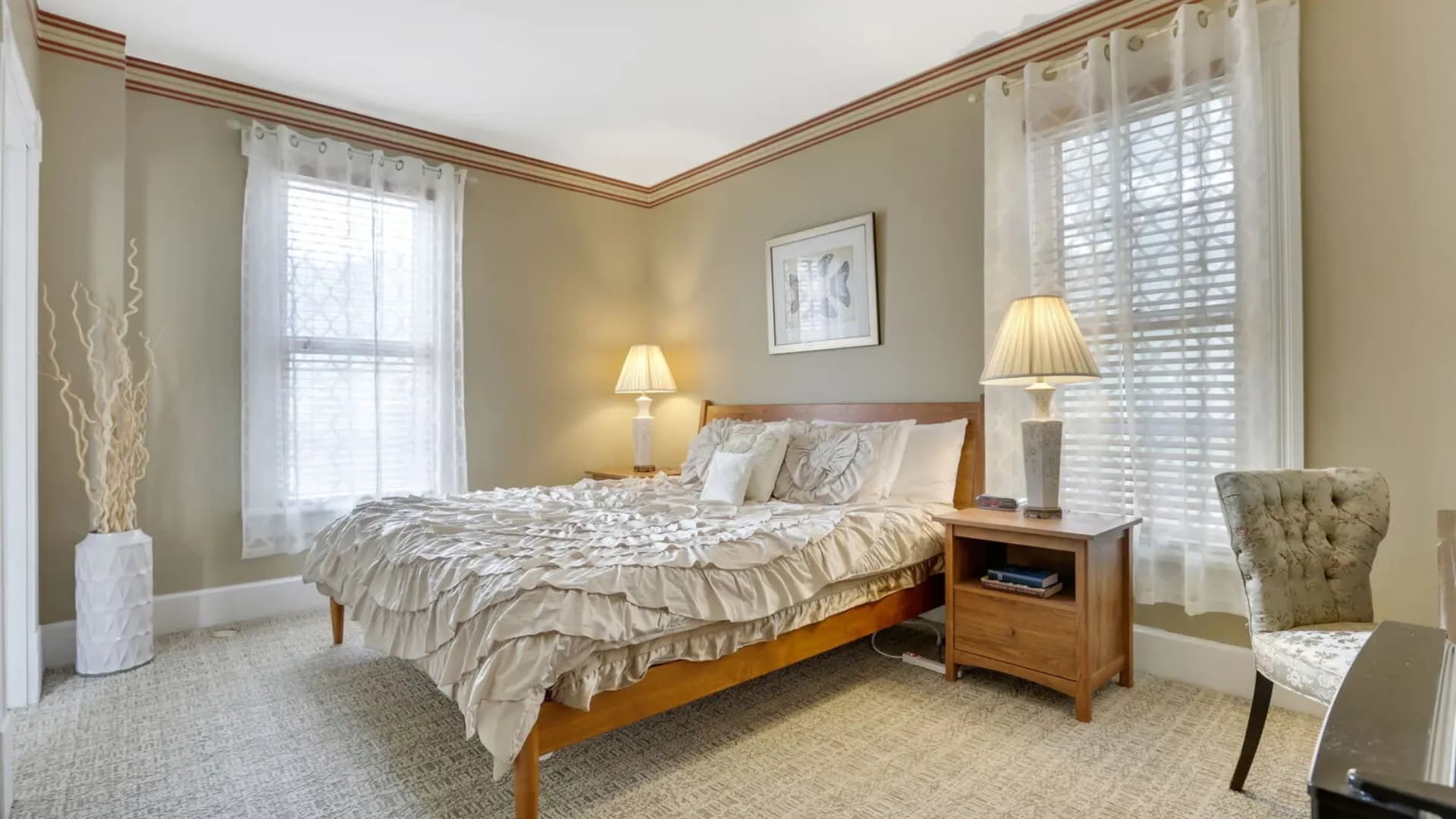 Cozy bedroom featuring a bed with ruffled bedding, bedside table, and large windows with sheer curtains.