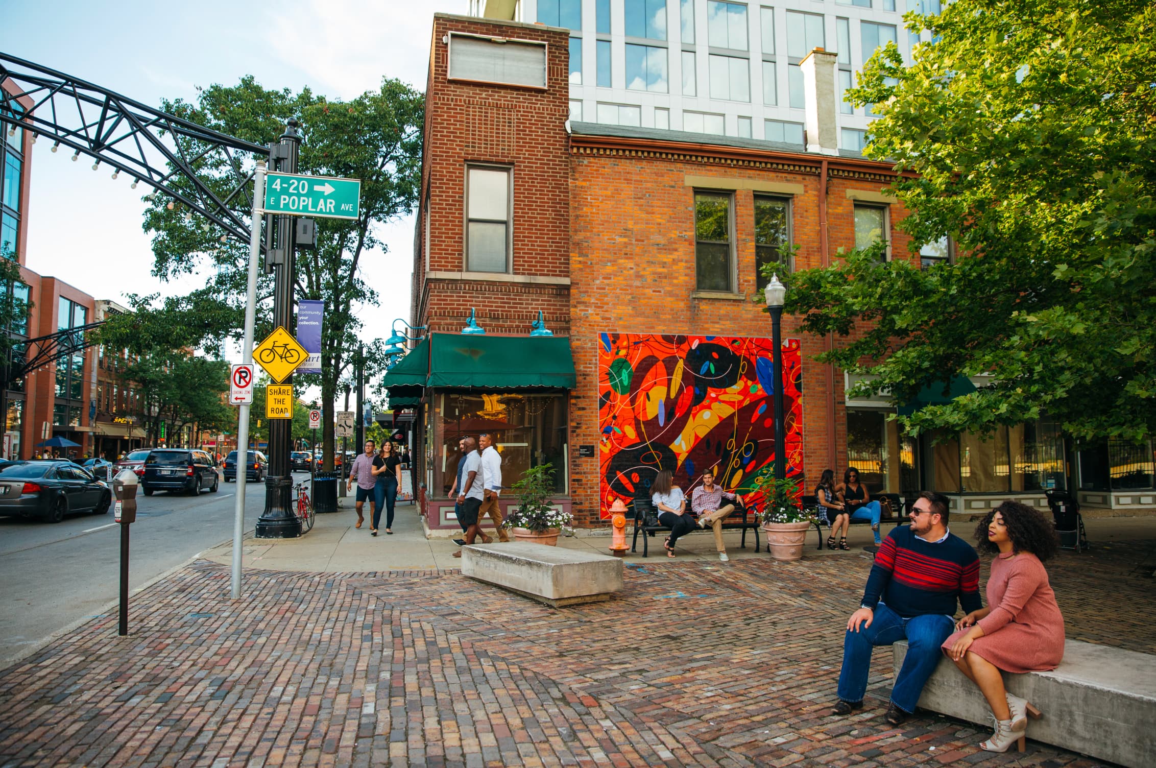 A busy street scene featuring a colorful mural, outdoor seating, and people walking in a vibrant urban area.